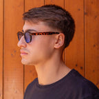 Model showcasing Deep Tortoise sunglasses with gradient blue lenses against a wooden background.
